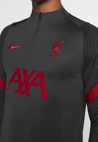 Mørkegrå træningstrøje med kvart lynlås, røde detaljer, Liverpool FC-logo og stort rødt "AXA"-tekst på fronten. Strukturerede sidelommer.