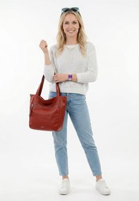 Smaidoša blondīne ar baltu džemperi, zilām džinsu biksēm, baltām sporta kurpēm, violetu pulksteni un lielu brūnu ādas rokassomu.