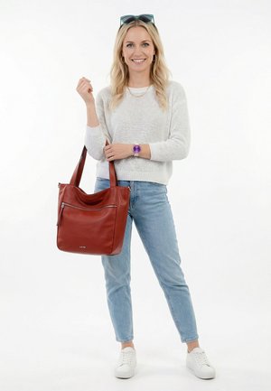 Smaidoša blondīne ar baltu džemperi, zilām džinsu biksēm, baltām sporta kurpēm, violetu pulksteni un lielu brūnu ādas rokassomu.