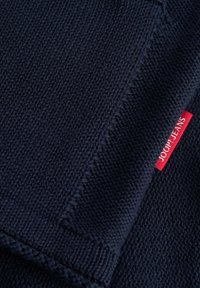 Tamsiai mėlynas megztas audinys su tekstūruotu raštu, krašte pritvirtinta maža raudona etiketė su užrašu „JOOP! JEANS“.