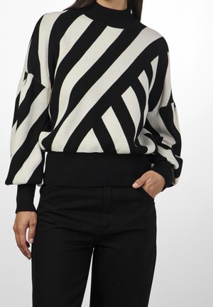 Persona con un suéter de rayas diagonales en blanco y negro con mangas abullonadas y pantalones negros, mano en el bolsillo.