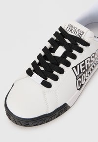 White sneaker with black laces, featuring "VERSACE JEANS COUTURE" text on the side and a black textured sole. Smooth material, low-top design.