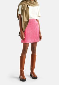 Woman wearing pink corduroy mini skirt, white short-sleeve top, light brown scarf, and knee-high brown leather boots standing on white background.