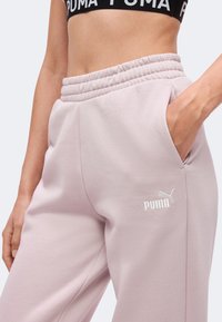 Γυναίκα που φοράει ανοιχτό ροζ φόρμα Puma με ελαστική μέση και πλαϊνές τσέπες, σε συνδυασμό με μαύρο αθλητικό μπουστάκι Puma.