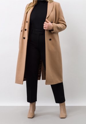 Kvinde iført en camel-farvet lang frakke over en sort rullekrave og sorte bukser, kombineret med beige ankelstøvler, stående mod en ensfarvet baggrund.