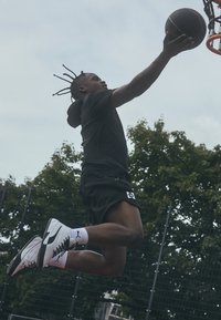 Atleta in pantaloni corti neri e felpa salta verso un canestro, indossando sneaker bianche e nere con accenti rosa, tenendo in mano un pallone da basket.
