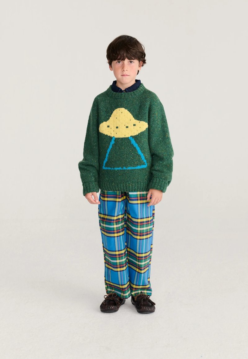 Garçon portant un pull vert avec un motif OVNI jaune, un pantalon à carreaux bleu et des mocassins foncés, debout devant un fond uni.