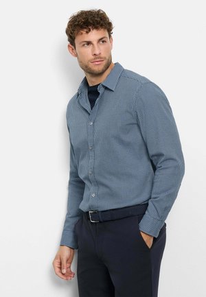 Man with curly hair wearing a blue small checkered button-up shirt, navy pants, and a black belt, looking to the side with one hand in pocket.