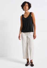Top negro sin mangas con tirantes texturizados, combinado con pantalones sueltos de color crema y zapatos negros trenzados, de pie contra un fondo blanco.