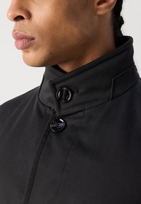 Zwarte jacket met een hoge kraag, voorzien van twee opvallende ronde knopen en een gladde stof textuur, die de op maat gemaakte ontwerpdetails benadrukt.
