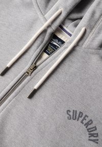 Grau zip-up Kapuzenpullover aus weichem Material, mit einer verstellbaren Kapuze, einem silbernen Reißverschluss und einem in Dunkelgrau bedruckten "SUPERDRY"-Logo.