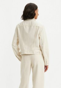 Chaqueta de mezclilla beige claro con un diseño ajustado, costuras visibles y dos botones en la parte trasera del dobladillo. Textura suave, estilo sencillo.