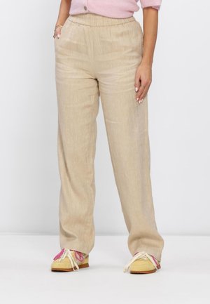 Vrouw draagt beige losse broek met elastische taille, een roze trui en gele sneakers met roze en witte veters, staand met de handen langs haar zij.
