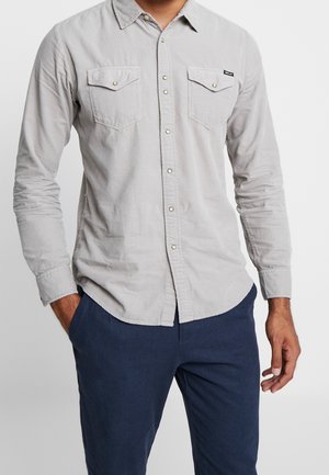 Hombre con camisa abotonada gris claro con bolsillos con solapas y pantalones azul marino, de pie con una mano en el bolsillo.