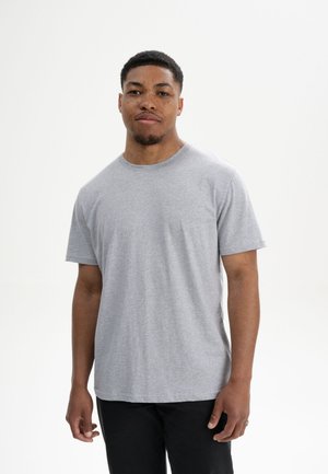 Jonge man met kort haar en sproeten, gekleed in een effen grijs t-shirt met korte mouwen en zwarte broek tegen een witte achtergrond.