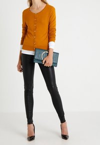 Senapsgul cardigan med knappar, lager på lager över en vit topp; glänsande svarta leggings; tealfärgad kuvertväska med en cirkulär detalj; svarta klackskor.