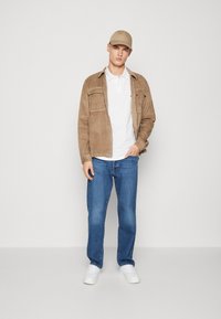 Beige kordflorsjacka över en vit polo, blå jeans och vita sneakers. Hatten matchar jackans färg, vilket skapar en koordinerad look.