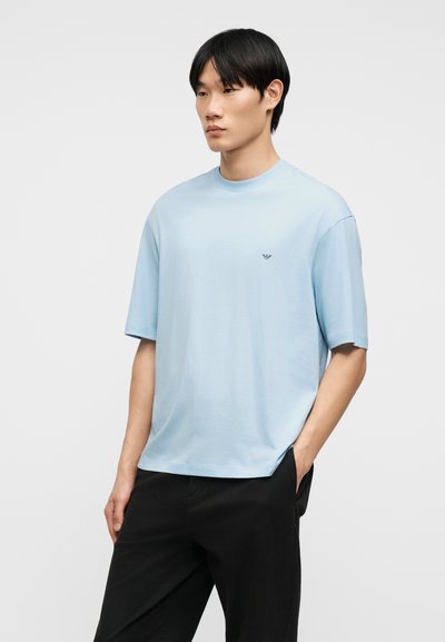 Νέος άνδρας που φοράει ανοιχτό μπλε κοντομάνικο πουκάμισο με μικρό λογότυπο στο στήθος και μαύρο παντελόνι, στέκεται με το ένα χέρι στην τσέπη.