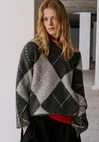 Pulover din lână cu model argyle în nuanțe de gri și negru, cu un design off-shoulder și manșete lănțuite, purtat peste un top roșu, având o textură moale.
