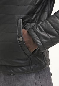 Giubbotto in pelle nera con motivo trapuntato, tasca laterale con chiusura a scatto e dettaglio testurizzato sulle maniche. Materiale robusto e design strutturato.