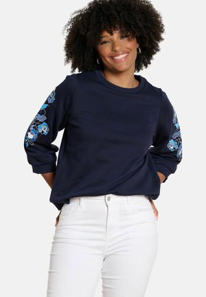Žena sa zakrútenými vlasmi, ktorá sa usmieva, oblečená v námornícky modrej mikine s modrým kvetinovým výšivkám na rukávoch a bielych nohaviciach, ruky vzadu za chrbtom.