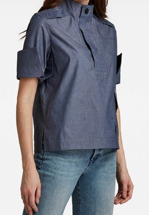 Chemise bleu foncé à manches courtes avec un col montant, une fermeture à boutons et des manches retroussées. Présente des coutures visibles et des fentes latérales.