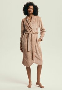 Woman with curly hair standing barefoot wearing a knee-length beige plush bathrobe with a belt tied at the waist and hands in pockets.