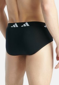 Schwarze Slips mit einem weißen adidas-Logo auf dem elastischen Bund. Glatte Textur, figurbetontes Design, das sich minimal am Körper anpasst.