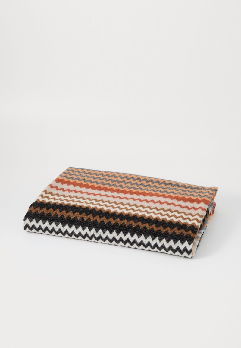 Tęczowy koc z wzorem w zygzaki, zawierający odcienie rdzy, kremu, czerni i brązu. Wykonany z miękkiego, teksturowanego materiału.