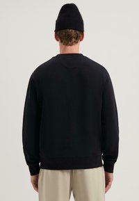 Czarny sweter z ribbowymi mankietami i dołem, o miękkiej fakturze, bez widocznych wzorów, noszony z kremowymi spodniami, widok z tyłu.