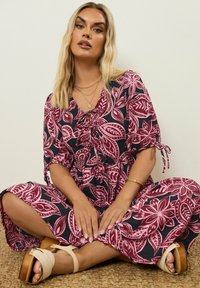 Blonde Frau, die im Schneidersitz auf einem geflochtenen Teppich sitzt, trägt ein lila Blumen-Kleid, goldene Armreifen, geschichtete Halsketten und beige High Heels.
