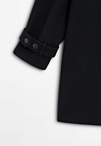 Manche d'un manteau noir avec une bride de poignet ajustable munie de deux boutons noirs, sur fond blanc.