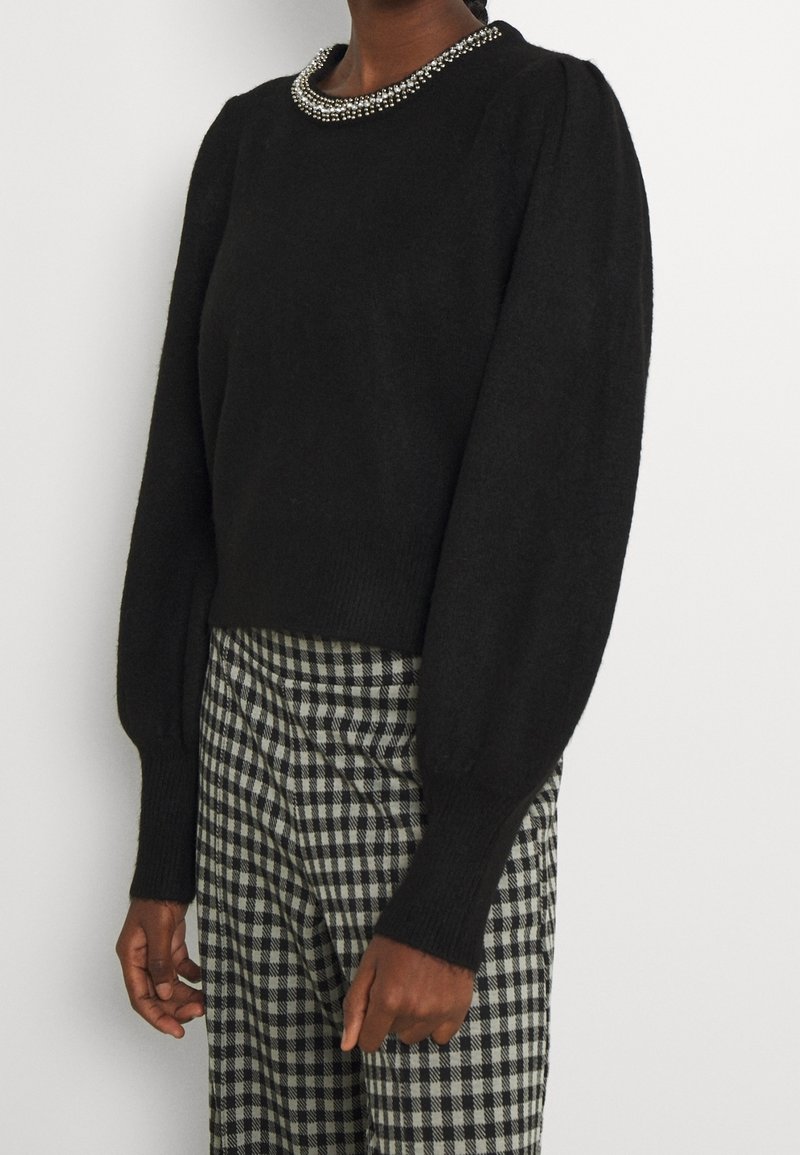 Femme portant un pull noir à manches longues avec un détail de col en perles et un pantalon taille haute à carreaux noirs et blancs.