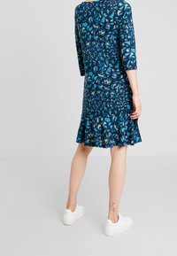 Blå och mönstrad klänning med trekvartsärmar, utsvängd nederkant, gjord av mjukt tyg; bärs med vita sneakers och har en ben-tatuering.