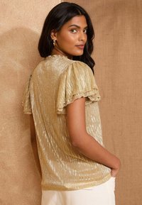 Gold metallic short-sleeve top with ruffled edges, textured finish, and a relaxed fit, worn with light-colored pants.