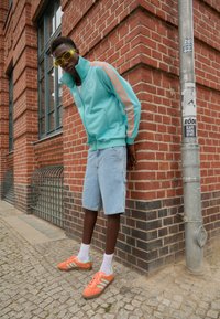 En turkis lynlåsjakke med orange striber, lyseblå denimshorts og orange sneakers med teal accenter, sat mod en murstensvæg.