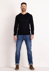 Maglione nero a maniche lunghe con scollatura a V abbinato a jeans azzurri chiari e scarpe marroni con stringhe. I jeans sono arrotolati alle caviglie.