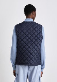 Navy quilted vest with a diamond pattern, soft material, sleeveless design, worn over a light blue long-sleeve shirt.