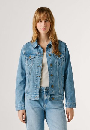 Jonge vrouw met lichtbruin haar, gekleed in een lichtblauwe spijkerjasje over een wit shirt en bijpassende spijkerbroek, staand tegen een effen achtergrond.