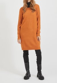 Oranges Strickkleid mit langen Ärmeln und gerippten Bündchen, kombiniert mit schwarzen, figurbetonten Hosen und schwarzen kniehohen Stiefeln. Einfaches und geradliniges Design.