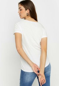 Valkoinen lyhytnuppinen T-paita, joka on valmistettu kevyestä materiaalista, rento malli ja pyöreä kaula-aukko, yhdistettynä sinisiin farkkuihin.