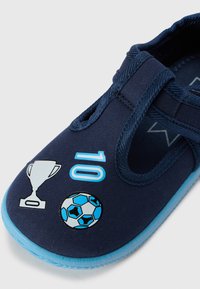 Chaussure à enfiler bleu marine avec semelle bleu clair. Présente des motifs sur le thème du football : un trophée, le numéro "10" et un ballon de football. Tissu texturé.