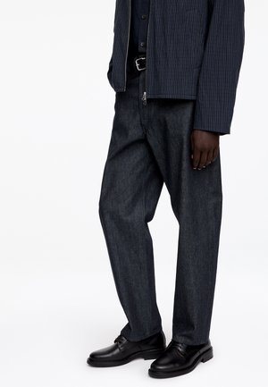 Mężczyzna ubrany w ciemną marynarkę w prążki, czarną koszulę, ciemne, luźne jeansy, czarny pasek i czarne skórzane eleganckie buty na białym tle.