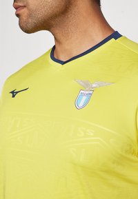 Lazio dzeltenā sporta krekls ar V veida kaklu, tumši zila apdare, teksturēts dizains un kluba logo ar spārnu simbolu uz krūšu.