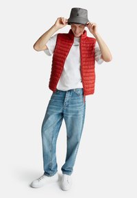 Chaleco acolchado rojo, camiseta blanca con texto, jeans sueltos de color azul claro y zapatillas blancas. Un gorro de pescador gris completa el conjunto.