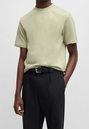 Man in een lichtbeige shirt met korte mouwen waarop het reliëflogo "HUGO" is gedrukt, en zwarte broek met een zwarte riem, met hand in zijn zak.