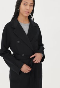 Svart dubbelknäppt kappa med stora knappar, bälte med knytband och vida ärmar. Bärs över en grå topp, med en jämn textur och skräddarsydd design.