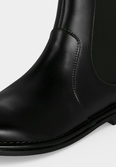 Gros plan d'une botte cheville en cuir noir avec panneau latéral élastique et coutures visibles le long de la semelle et de la tige.