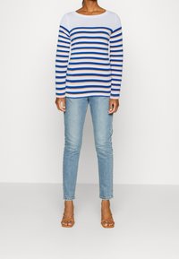 Wzorzysty sweter z długim rękawem w białym kolorze z niebieskimi i pomarańczowymi paskami, zestawiony z jasnoniebieskimi dżinsami i brązowymi sandałami na paski.