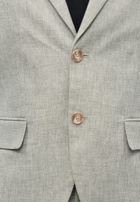 Φωτεινό γκρι υφασμάτινο σακάκι με δύο καφέ κουμπιά κλειστά και τσέπες με καπάκια και στις δύο πλευρές, φορεμένο πάνω από μαύρο τοπ.